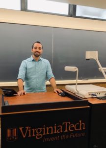 Tim Beck stands at the front of his classroom with the words "Virginia Tech: Invent the Future" printed on the front of his desk