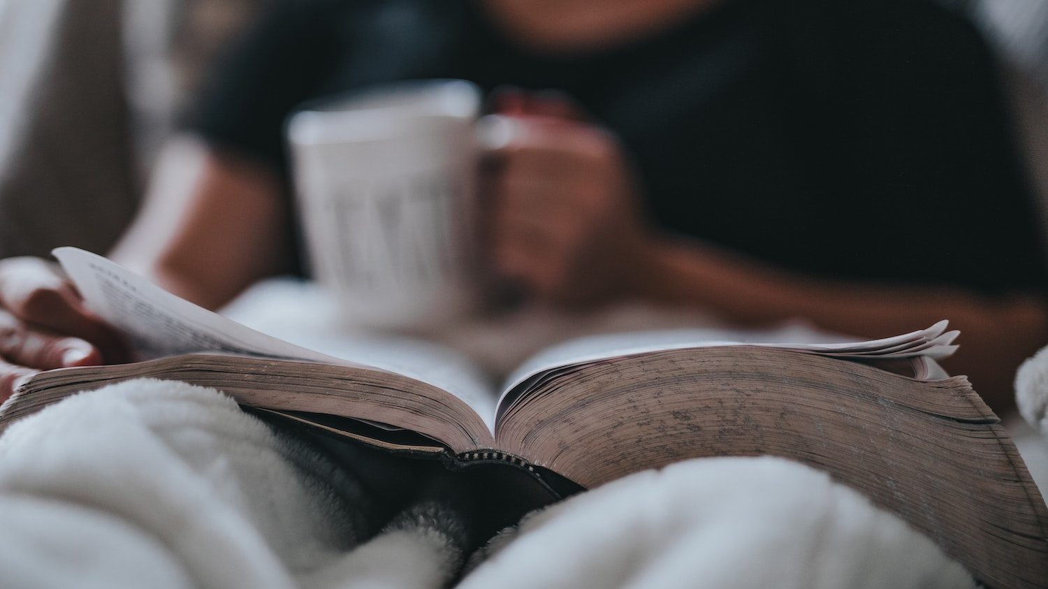 someone holding a mug and a book while lying in bed