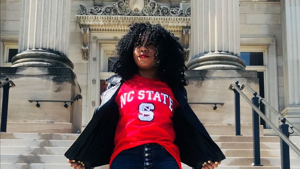 Chelsea Smith wearing a red NC State t-shirt and jeans, standing in front of a building and looking into distance