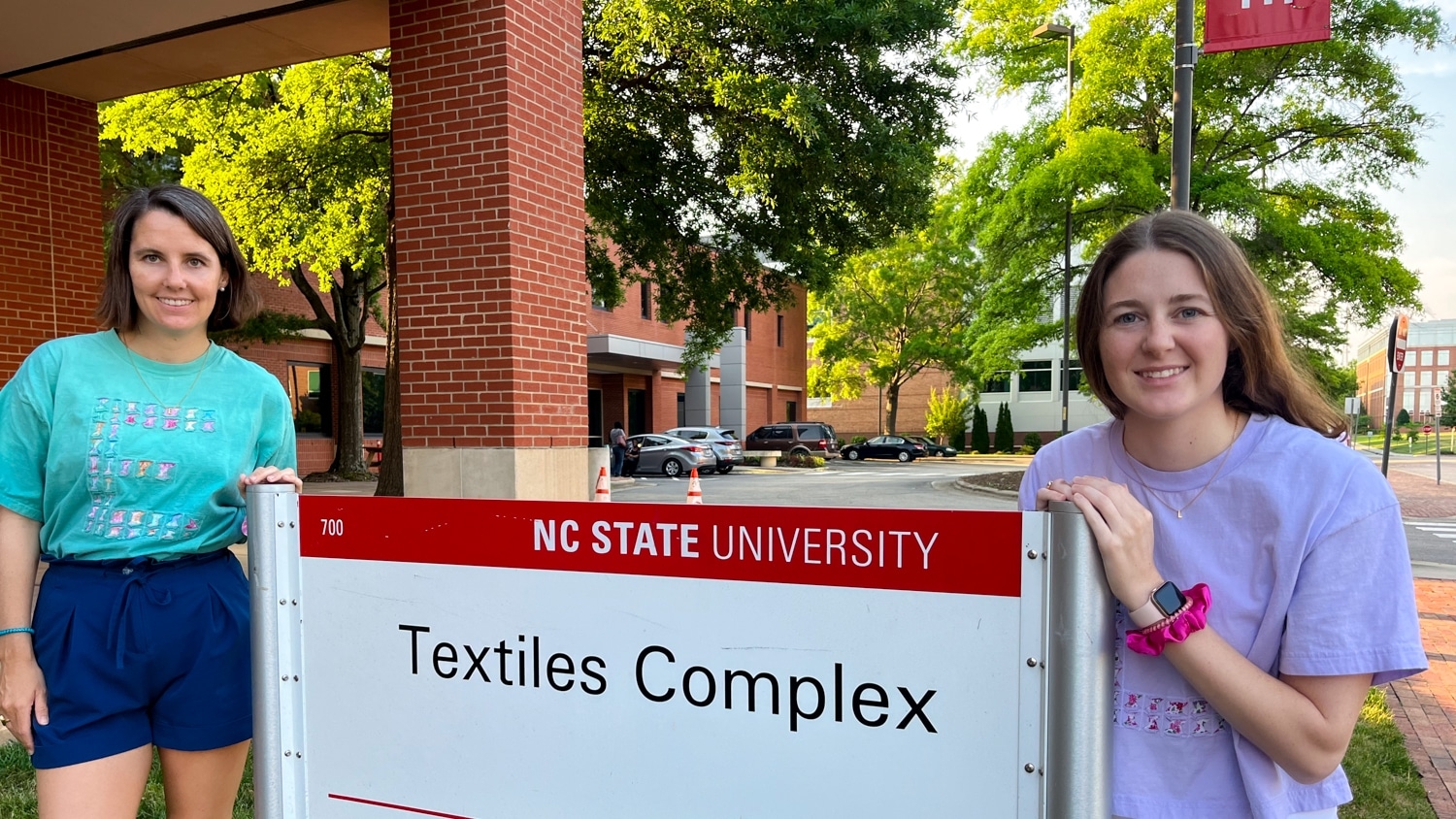 Emily Potok (left) and Laura Potok (right) stand on either side of a white sign that reads "Textiles Complex" at the Wilson College of Textiles campus.