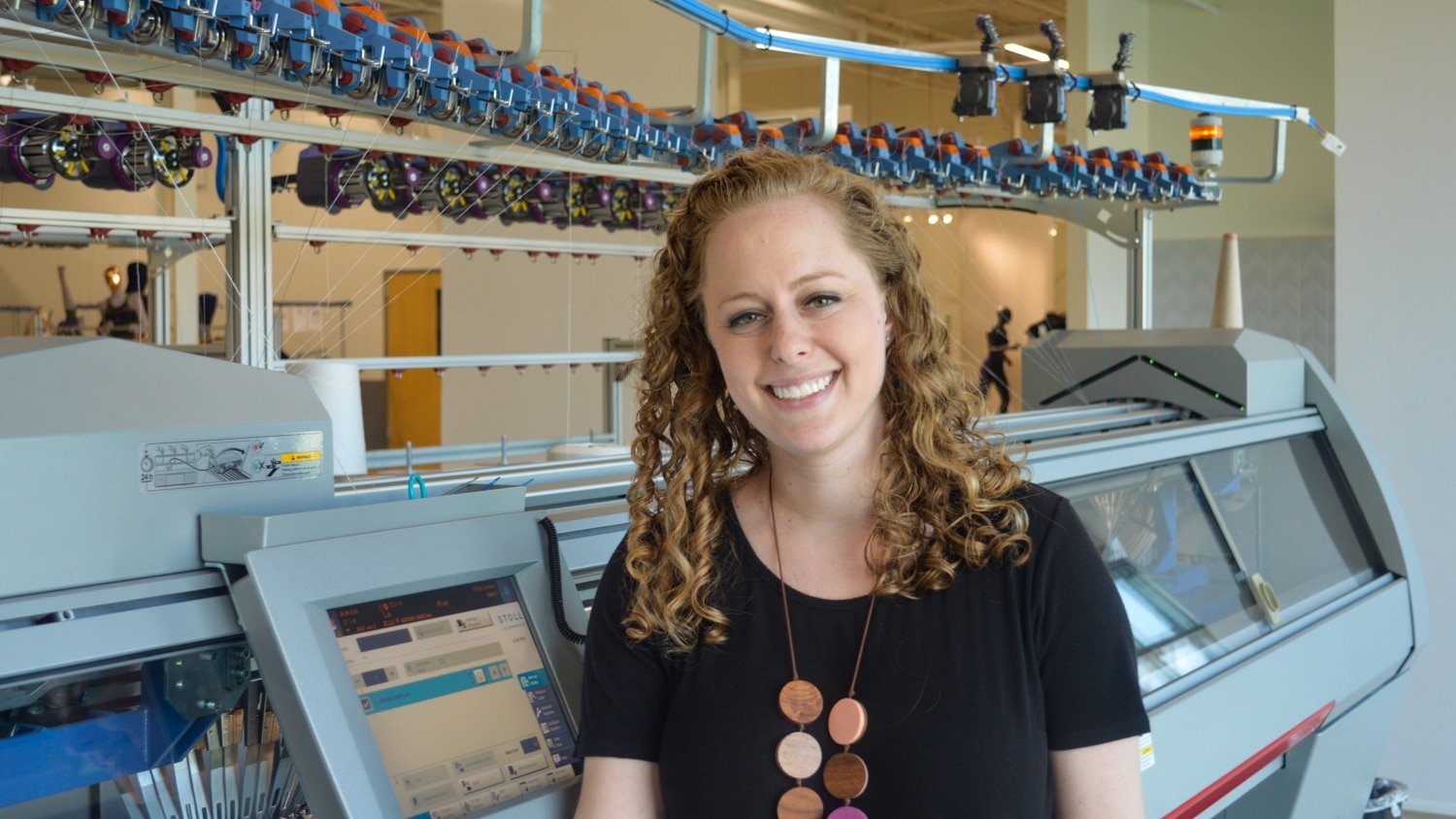 Zoe Newman stands in front of a knitting machine and smiles at the camera.