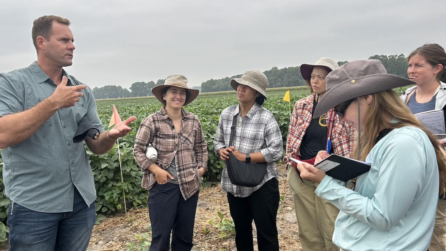 Students gather round a man in a farm field and take notes.